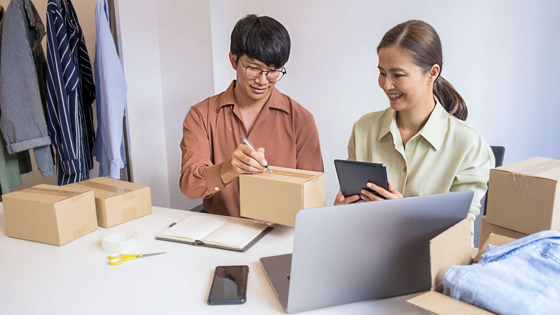 Deux entrepreneurs de PME préparant des colis et gérant leurs commandes en ligne à l’aide d’un ordinateur et d’une tablette, illustrant une gestion moderne et organisée